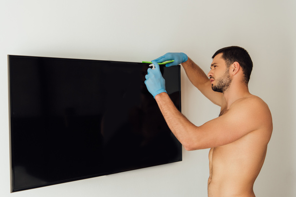 shirtless man in rubber gloves holding rag and bottle with antibacterial liquid near tv with blank screen - Photo, Image