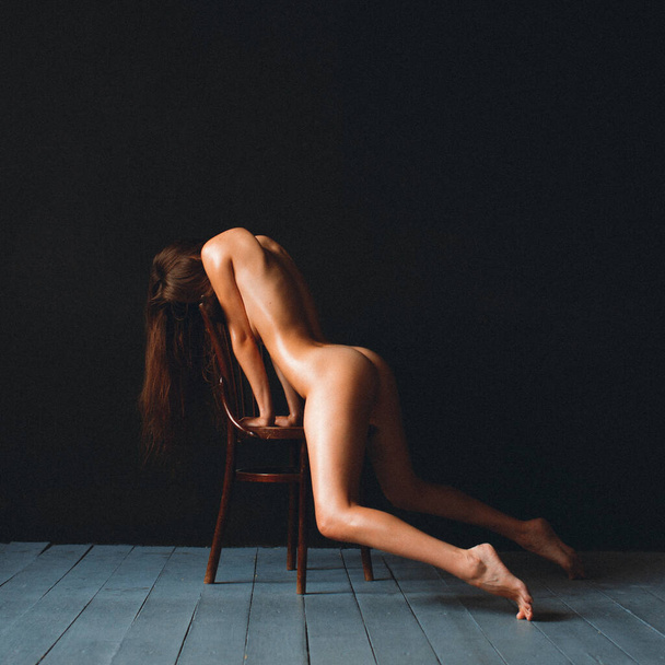 Young beautiful girl with good body shapes, nude, against a black wall, sits on an old chair, her body is rubbed with oil - Fotó, kép