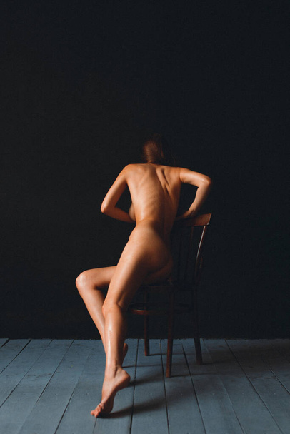 Young beautiful girl with good body shapes, nude, against a black wall, sits on an old chair, her body is rubbed with oil - Fotografie, Obrázek
