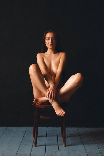 Junge schöne Mädchen mit guten Körperformen, nackt, vor einer schwarzen Wand, sitzt auf einem alten Stuhl, ihr Körper ist mit Öl eingerieben - Foto, Bild