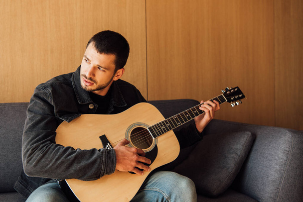 parrakas mies soittaa akustista kitaraa olohuoneessa
  - Valokuva, kuva