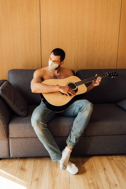 ημίγυμνος άνδρας με ιατρική μάσκα που παίζει ακουστική κιθάρα στο σαλόνι  - Φωτογραφία, εικόνα