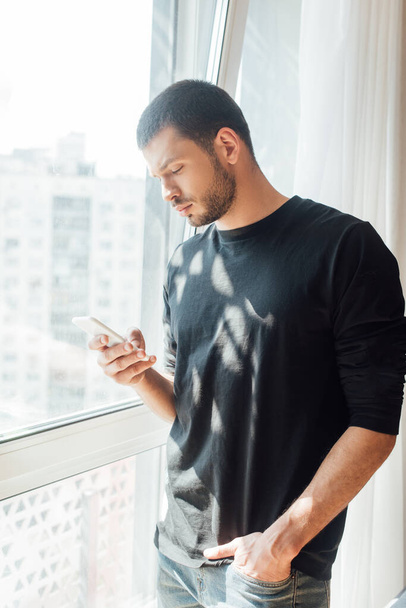 uomo barbuto in piedi con la mano in tasca e utilizzando smartphone vicino alla finestra a casa
  - Foto, immagini