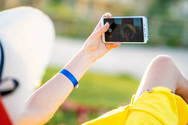 Νεαρή γυναίκα με κίτρινο καλοκαιρινό φόρεμα που βγάζει selfie με το κινητό της τηλέφωνο ακουμπισμένη σε παγκάκι στο πάρκο. - Φωτογραφία, εικόνα