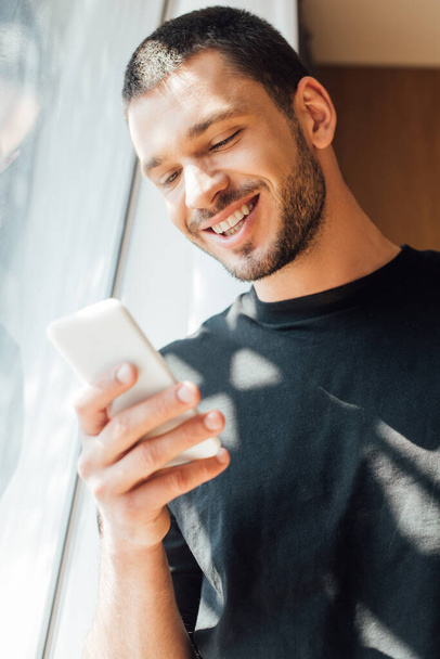 χαμηλή γωνία άποψη του χαρούμενος άνθρωπος χρησιμοποιώντας smartphone κοντά στο παράθυρο στο σπίτι  - Φωτογραφία, εικόνα
