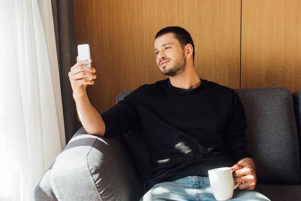 luz del sol en el hombre guapo sonriendo mientras usa el teléfono inteligente y sostiene la taza en la sala de estar
  - Foto, imagen