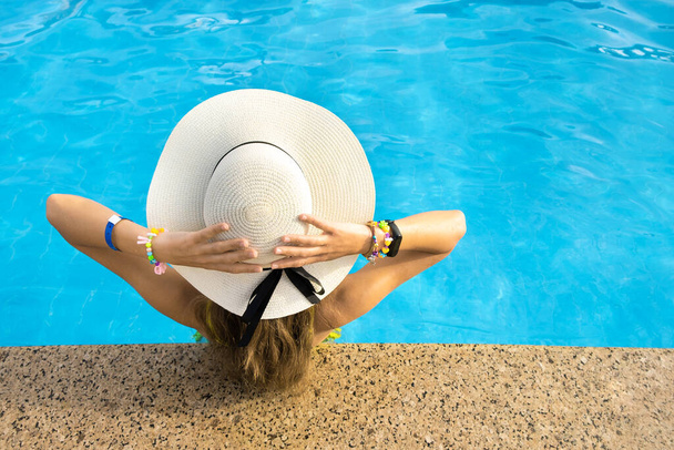 長い髪をした若い女性の背中のビュー晴れた日に青い水で暖かい夏のスイミングプールでリラックス黄色のわら帽子を身に着けています. - 写真・画像