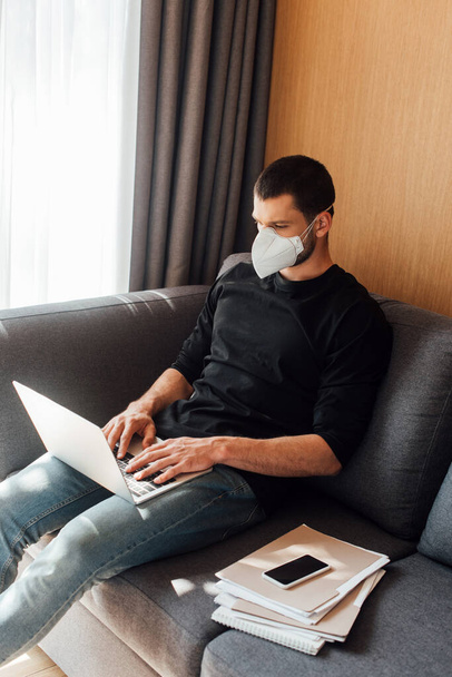 blogger en máscara médica usando laptop cerca de smartphone con pantalla en blanco en casa
 - Foto, imagen