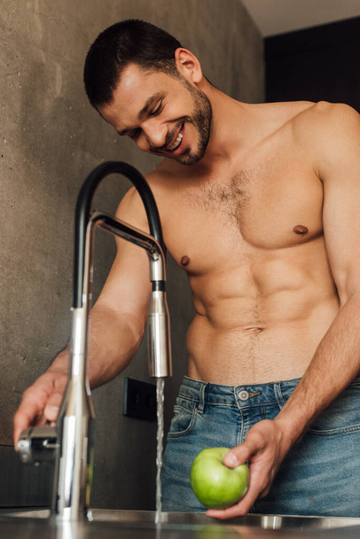 selective focus of shirtless man smiling while holding apple with faucet with pouring water in kitchen  - Valokuva, kuva