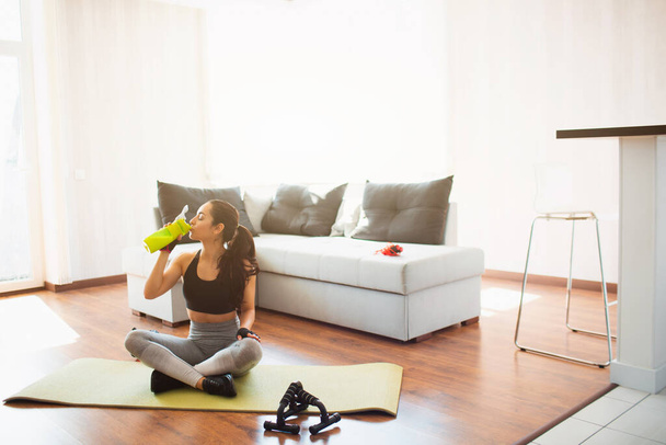 Young woman doing sport workout in room during quarantine. Sit on mat with crossed legs and drink protein from green bottle. Rest relax after workout. - Photo, Image