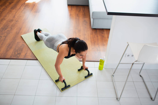 Young woman doing sport workout in room during quarantine. Up side view of girl stand in plank position using push up stands hand bar. Exercising in room. - Photo, Image