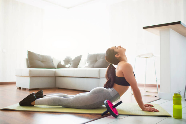 Mujer joven haciendo ejercicio deportivo en la habitación durante la cuarentena. Retrocediendo en la esterilla de yoga. Acostado sobre el estómago y estirándose
. - Foto, imagen