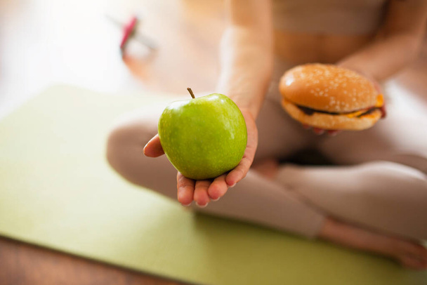 Mujer joven haciendo ejercicios de yoga en la habitación durante la cuarentena. Corte la vista de la niña sosteniendo manzana verde saludable y hamburguesa gorda en las manos. Es una decisión difícil. Estilo de vida saludable o poco saludable
. - Foto, imagen