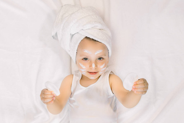 Pretty 2 years old girl child relaxing in the bedroom after shower, lying on the bed, wearing white towel on head and holding in hands two cotton pads. Morning care and baby hygiene - 写真・画像