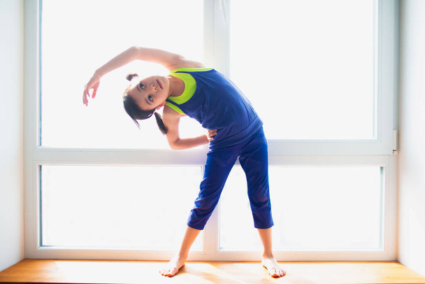 Маленькая девочка делает задний наклон стоя тренировки дома. Милый парень тренируется на деревянном подоконнике в помещении. Маленькая темноволосая модель в спортивной одежде имеет упражнения возле окна в своей комнате
 - Фото, изображение