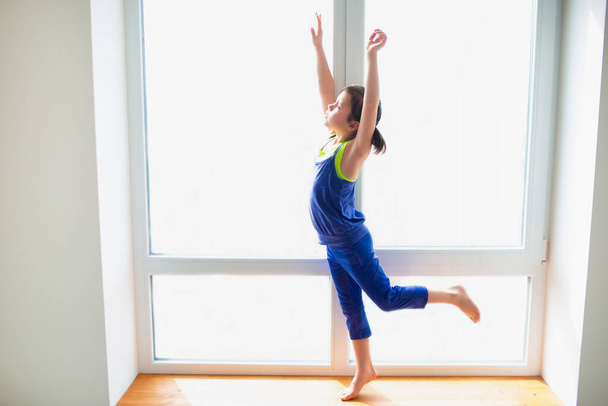 Little girl reaching up workout at home. Cute kid is training on a wooden windowsill indoor. Little dark-haired female model in sportswear has exercises near the window in her room. - Photo, Image