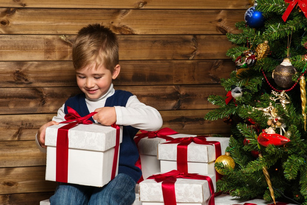 kleine jongen openen geschenkdoos onder de kerstboom in houten huis interieur - Foto, afbeelding