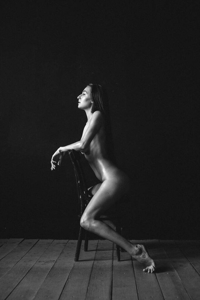 Young beautiful girl with good body shapes, nude, against a black wall, sits on an old chair, her body is rubbed with oil - Foto, imagen