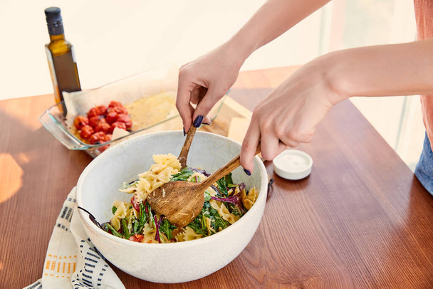 Cropped view of woman mixing pasta salad in bowl with spatulas near napkin at table in kitchen - Foto, Imagem