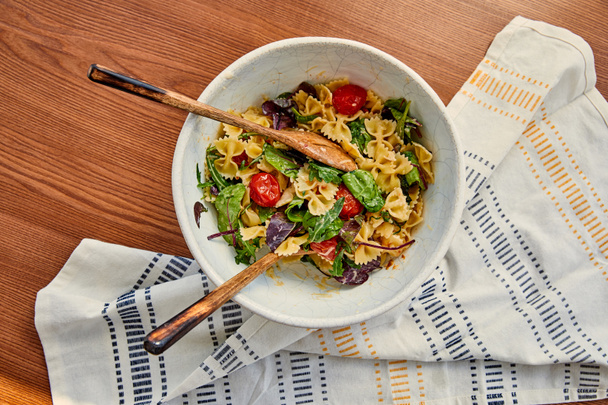 Top view of bowl with pasta salad and spatulas near napkin on wooden background - Valokuva, kuva