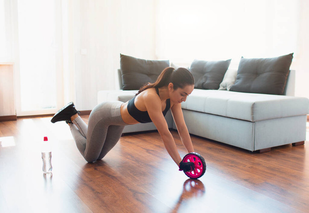 Young woman doing sport workout in room during quarantine. Stand on knees and using abdominal exercise roller for body stretching. Home training. - Photo, Image