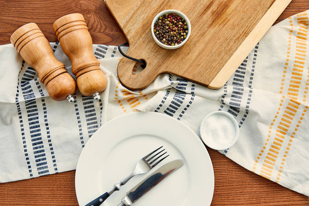 Top view of plate with fork and knife near napkin, cutting board, bowls, pepper and salt mills on wooden background - Foto, Imagem