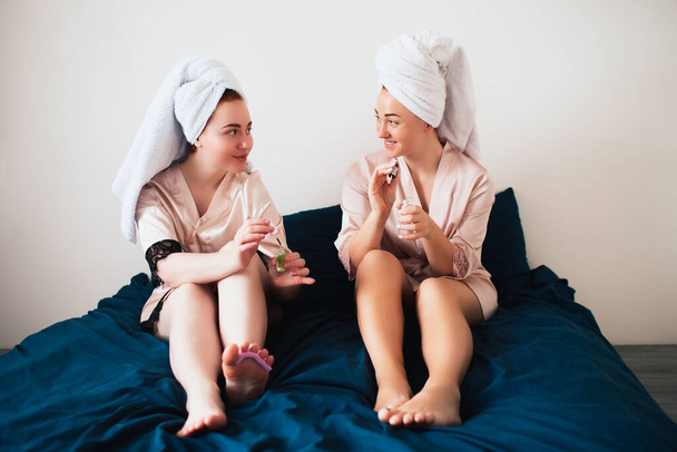 Dwie młode kobiety w ręcznikach i piżamie urządzają w domu zabawną imprezę spa. Siedzą na łóżku i robią sobie nowy pedicure. - Zdjęcie, obraz