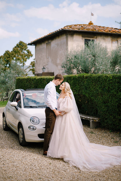 Schöne Braut und Bräutigam schauen einander an und halten Händchen vor einem Cabrio in der Villa in der Toskana, Italien - Foto, Bild