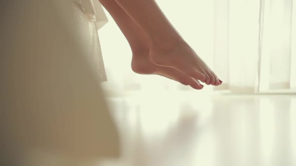close-up shooting. legs of a pretty girl getting out of bed against the window. light background, morning wakes up. white bed beautiful female toes. go on the floor. parquet or laminate. fitness legs  - Felvétel, videó