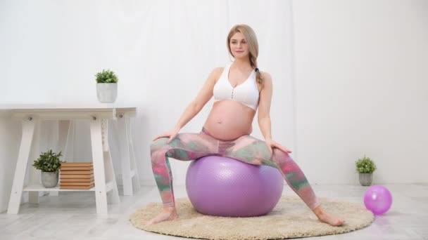 A young beautiful pregnant Caucasian woman sits on the soft carpet of the house. He is engaged in fitness and doing exercises. Sportswear. - Imágenes, Vídeo