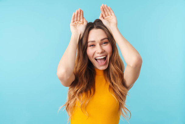 Πορτρέτο της όμορφης χαρούμενης γυναίκας χαμογελώντας και gesturing αυτιά λαγουδάκι με τα χέρια απομονώνονται πάνω από το μπλε φόντο στο στούντιο - Φωτογραφία, εικόνα