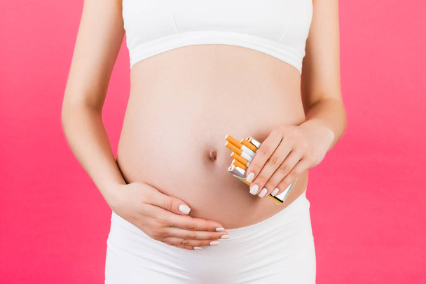 Close up de mulher grávida de roupa interior branca segurando um maço de cigarros em fundo rosa. Estilo de vida pouco saudável durante a gravidez. Dependência tabágica
. - Foto, Imagem