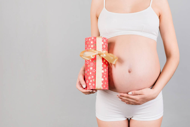 Beyaz iç çamaşırlı hamile bir kadının hediye kutusunu tutarken ve mavi arka planda karnına dokunurken görüntüsü kesilmiş. Bir kız bebek bekliyorum. Boşluğu kopyala. - Fotoğraf, Görsel