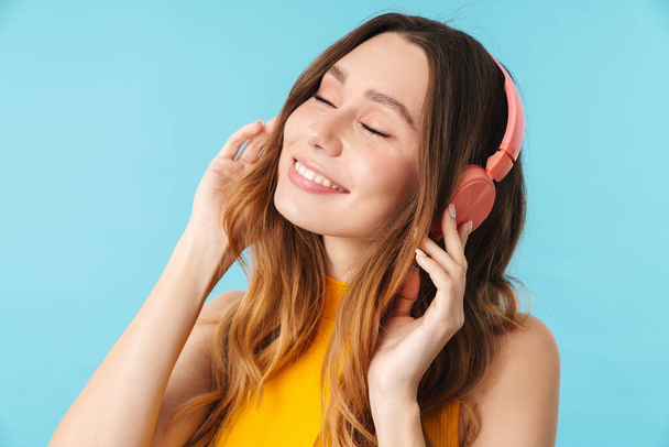 Portrait of beautiful joyous woman wearing headphones listening to music isolated over blue background in studio - Photo, Image