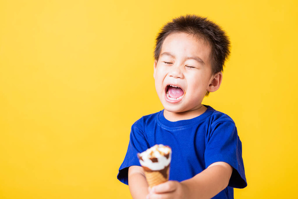 Happy portrait Asian child or kid cute little boy attractive laugh smile playing holds and eating sweet chocolate ice cream waffle cone, studio shot isolated on yellow background, summer concept - Foto, afbeelding
