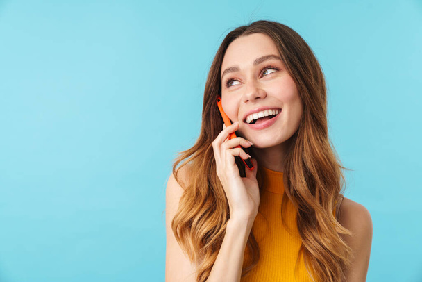 Portrait of beautiful joyous caucasian woman smiling and talking on cellphone isolated over blue background in studio - Фото, зображення