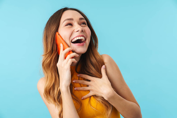 Portrait of beautiful joyous caucasian woman smiling and talking on cellphone isolated over blue background in studio - Fotografie, Obrázek