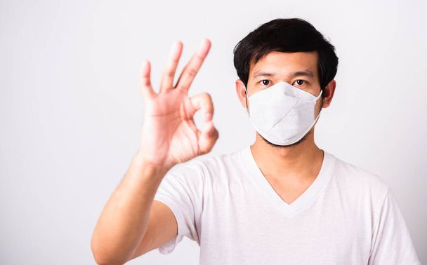 Closeup Asian handsome Man wearing surgical hygienic protective cloth face mask against coronavirus and show OK sign, studio shot isolated white background, COVID-19 medical concept - Foto, Bild