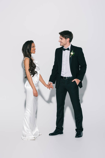 full length view of happy, elegant interracial newlyweds holding hands and looking at each other on white background - Фото, изображение