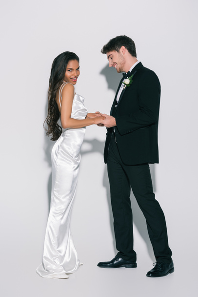 full length view of happy, attractive african american bride looking at camera while holding hands with elegant groom on white background - Fotografie, Obrázek