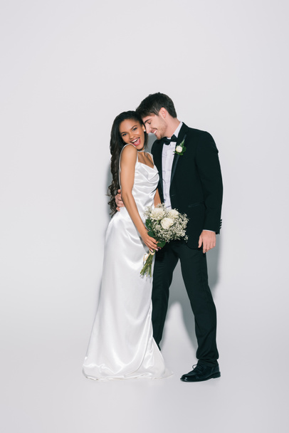 plná délka pohled na šťastný ženich objímání elegantní africký americký nevěsta drží svatební kytice na bílém pozadí - Fotografie, Obrázek