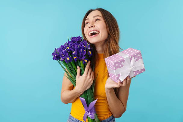 Portrait of joyous young happy woman smiling while holding flowers and gift box isolated over blue background in studio - Foto, imagen