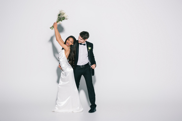 full length view of happy african american bride holding wedding bouquet in raised hands near elegant bridegroom on white background - Photo, Image