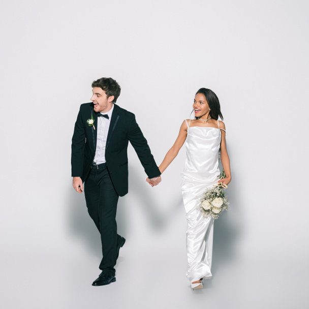 full length view of smiling, elegant interracial newlyweds holding hands while walking and looking away on white background - Foto, Bild
