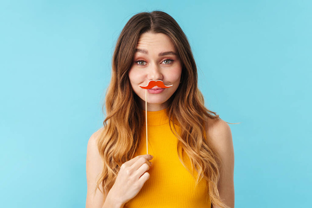 Portrait of a cheerful pretty young girl standing isolated over blue background, wearing fake moustache - Photo, Image