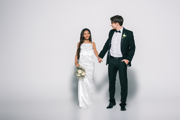 full length view of handsome bridegroom with hand in pocket holding hands with beautiful african american bride on white background - Fotografie, Obrázek