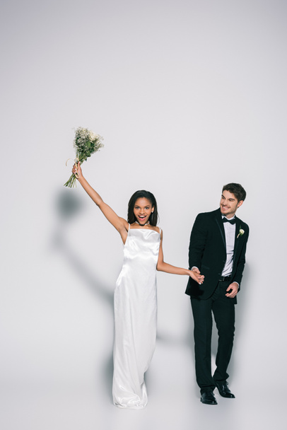 full length view of happy african american bride holding wedding bouquet in raised hand while holding hands with elegant bridegroom on white background - Photo, Image