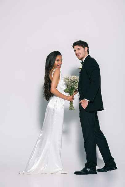 full length άποψη του χαμογελαστού γαμπρού νύφη κρατώντας το χέρι στην τσέπη και κοιτάζοντας μακριά κοντά στην αφρικανική αμερικανική νύφη κοιτάζοντας κάμερα σε λευκό φόντο - Φωτογραφία, εικόνα