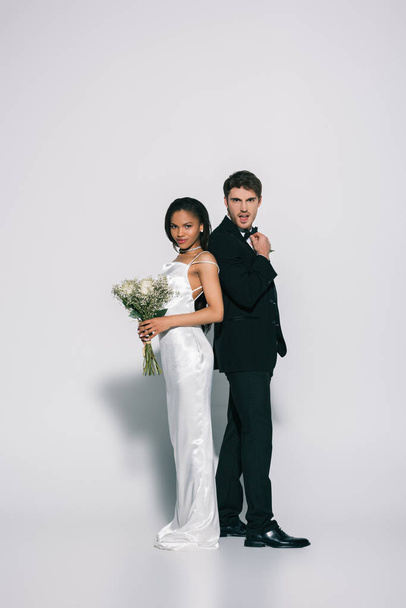 full length view of young, elegant interracial newlyweds standing back to back while posing on white background - Фото, изображение