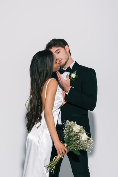 young bridegroom touching face and kissing beautiful african american bride on white background - Zdjęcie, obraz